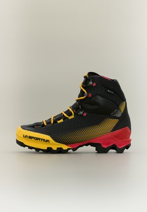Vysoká turistická obuv v čiernej farbe so žltými a červenými akcentmi, hrubou podrážkou, žltými šnúrkami a značkou "La Sportiva" na oblasti priehlavku.