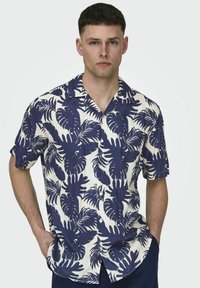 Camicia a maniche corte color crema con un motivo di palme blu e foglie, dotata di abbottonatura frontale e colletto classico.
