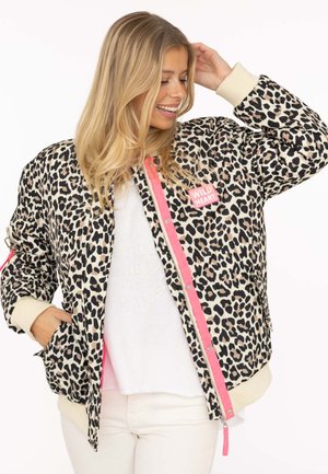 Lächelnde Frau mit langen blonden Haaren, die eine Jacke mit Leopardenmuster und rosa Reißverschluss sowie einen "Wild Heart"-Aufnäher, ein weißes Hemd und eine weiße Hose trägt.