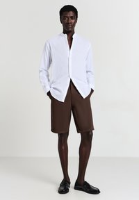 Homme portant une chemise blanche à col mandarin, un short marron à hauteur de genoux et des mocassins noirs, se tenant les mains dans les poches sur un fond uni.