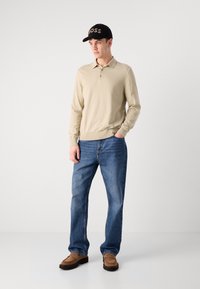 Polo shirt beige a maniche lunghe, jeans blu, scarpe marroni e un cappellino nero con il logo "BOSS". Il materiale sembra morbido con una vestibilità rilassata.