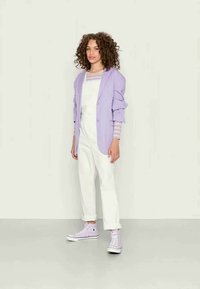Blazer lavanda sobre um macacão branco com punhos enrolados. Ténis lavanda com solas brancas. Textura suave, estilo casual, corte contemporâneo.