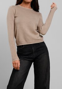 Maglione beige a maglia con scollo rotondo, polsini e vita a coste. Abbinato a pantaloni in denim scuri per un look casual.