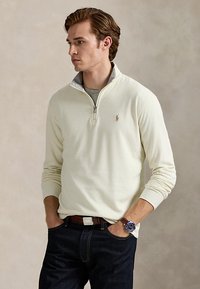 Junger Mann mit Händen in den Taschen, trägt einen elfenbeinfarbenen Viertel-Reißverschluss-Pullover, dunkle Jeans, braunen Gürtel und eine Uhr mit blauem Zifferblatt.