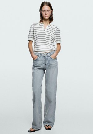 Kobieta w białej i szarej koszulce polo w paski na krótki rękaw, jasnobłękitnych szerokich dżinsach i czarnych sandałach, z rękami w kieszeniach.