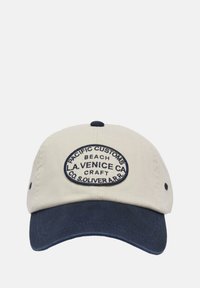 Beige katoenen pet met een marineblauwe rand. Bevat een ovale patch met geborduurde tekst: "PACIFIC CUSTOMS BEACH LA VENICE CA CRAFT CO. S.OLIVER."