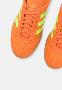 Orange suede sneakers featuring neon green and white stripes, flat laces, and a gum sole. Adidas logo on tongue and side.