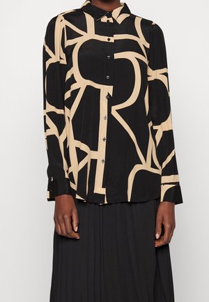 Femme portant un chemisier noir avec des motifs abstraits de lignes beiges et des boutons noirs, associé à une longue jupe noire.