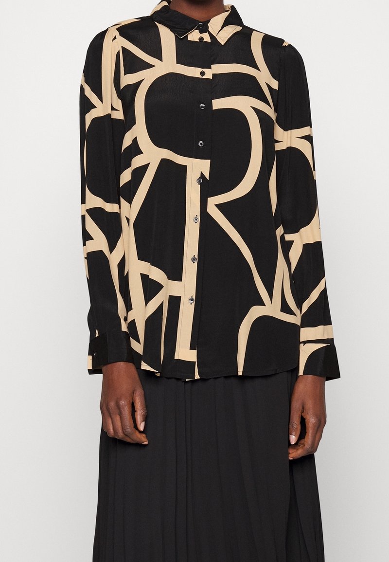 Femme portant un chemisier noir avec des motifs abstraits de lignes beiges et des boutons noirs, associé à une longue jupe noire.