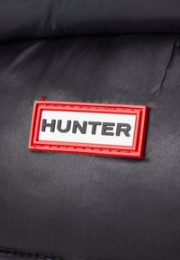 Tejido negro con una etiqueta rectangular en rojo y blanco que lleva la palabra "HUNTER" en relieve. Textura suave, diseño minimalista, logo prominente.