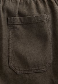 Dark brown fabric with a textured surface, featuring a square patch pocket and elastic waistband. Stitching is visible around the edges.