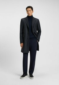 Cappotto di lana scuro con motivo a scacchi, tre bottoni, abbinato a un dolcevita blu navy e pantaloni blu navy su misura. Sneakers nere completano il look.