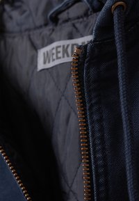 Námořnická modrá bunda s prošívanou podšívkou. Obsahuje výrazný zip v bronzovém tónu a štítek s nápisem "WEEKEND." Hladká textura.