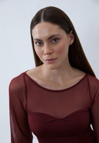 Bordó hosszú ujjú felső áttetsző hálós ujjal, testhez simuló fazonnal és puha, kerek dekoltázzsal. Sima textúra, finom fényességgel.
