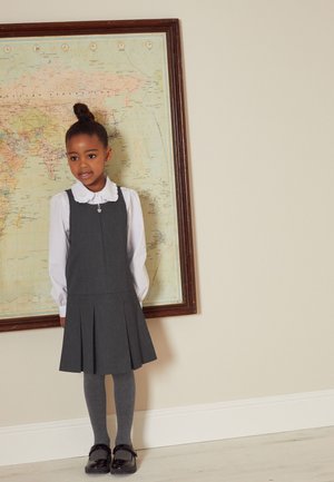 Graues Latzkleid mit Falten, getragen über einem weißen Hemd mit Kragen. Graue Strumpfhosen und schwarze Schuhe. Der Hintergrund zeigt eine Karte in einem Holzrahmen.