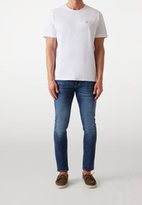 Hombre vestido con una camiseta blanca de manga corta, jeans ajustados azules y zapatos casuales marrones con cordones, de pie contra un fondo blanco.