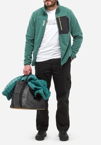 Veste en polaire verte avec panneau noir et zip jaune, associée à un pantalon cargo noir. Tenant une veste rembourrée teal dans un tote bag noir.