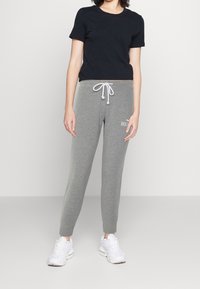 Kvinna klädd i en svart kortärmad topp, grå joggingsbyxor med dragsko och logotyp, samt vita sneakers stående framför en enfärgad bakgrund.