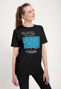 Mladá žena s culíkem, oblečená v čern�ém tričku s Picasso dílem „Tři tanečnice“, které obsahuje modrý a bílý text.