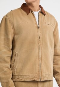 Giacca zip-up color cammello con colletto in velluto a coste marrone, tasca frontale e leggeri dettagli di cucitura. Realizzata in un tessuto durevole con una trama liscia.