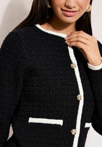 Naine, kes kannab tekstuuriga musta kardigani, millel on valged ääred ja kuldsed lillekujulised nööbid, puudutab krae õla lähedal.