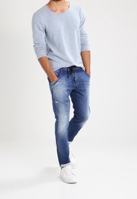 Hombre con una camiseta de manga larga gris claro, jeans azules desgastados con cordón, manos en los bolsillos y zapatillas blancas frente a un fondo liso.