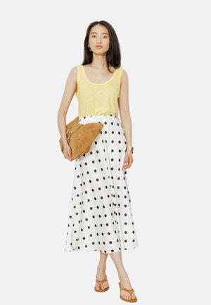 Femme portant un débardeur jaune, une jupe midi blanche à pois noirs, des sandales marron clair, tenant une grande pochette en suède marron clair.