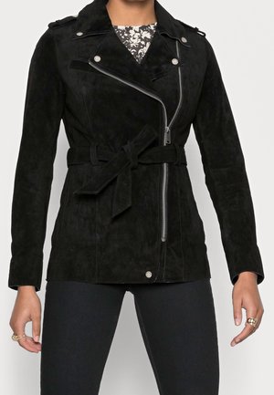 Personne portant une veste en suède noire ceinturée avec une fermeture éclair argentée asymétrique et un haut floral noir et blanc en dessous.