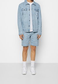 Lichtblauwe denimjacket met een patroon, gecombineerd met bijpassende shorts. Witte t-shirt eronder, gedragen met witte sneakers en sokken.