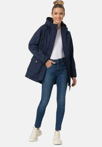 Navy waterproof jacket with a hood, zip and snap closure, and multiple pockets. Paired with blue jeans and white sneakers.