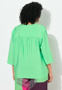 Blusa verde con una vestibilità comoda, maniche a tre quarti e un dettaglio plissettato sul retro, realizzata in tessuto leggero e texturizzato.