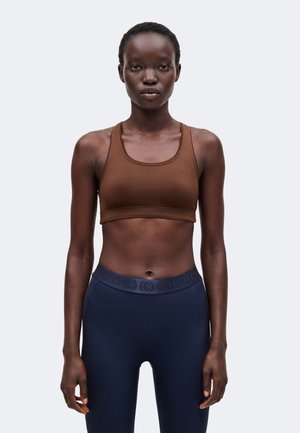 Kvinde stående forfra iført en brun sports-bh og mørkeblå leggings med høj talje mod en ensartet lys baggrund.