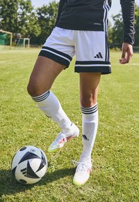 Weiße Fußballshorts mit schwarzen Streifen, Adidas-Logo, kombiniert mit weißen Socken und Fußballschuhen. Ein schwarz-weißer Fußball liegt daneben.