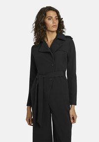 Zwarte trenchcoat met lange mouwen, grote revers, dubbele knopen en een gestrikte taille. Glad textuur en op maat gemaakte pasvorm.