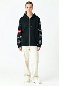 Felpa nera con zip e stampe grafiche in bianco e accenti colorati, abbinata a pantaloni cargo crema e sneakers bianche.