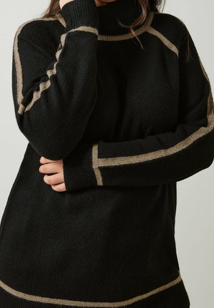 Femme portant un pull à col roulé noir avec des rayures beiges le long des manches, des épaules et de l'ourlet, les bras croisés de manière décontractée.