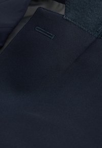 Tela azul marino con una textura suave y un ligero brillo, que presenta un acento cosido y un pequeño detalle en bucle horizontal.