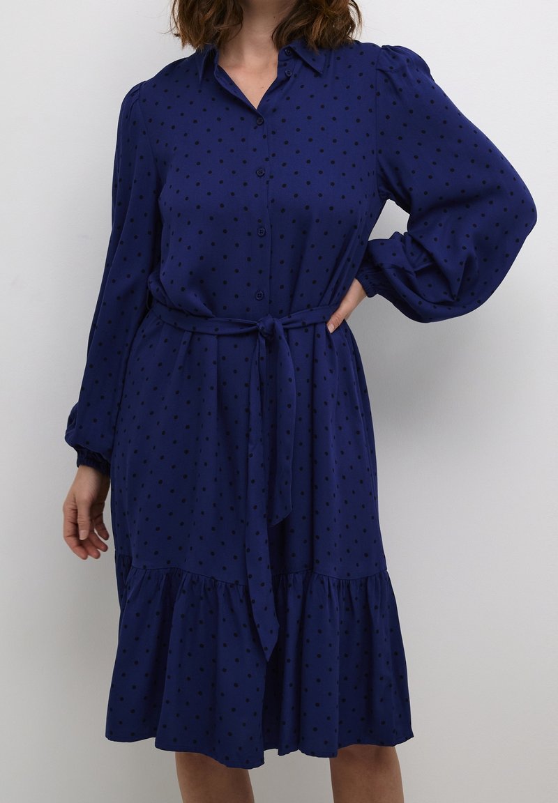 Femme portant une robe bleu marine à manches longues avec des pois noirs, un col, des boutons sur le devant, une ceinture nouée à la taille et un ourlet à volants.