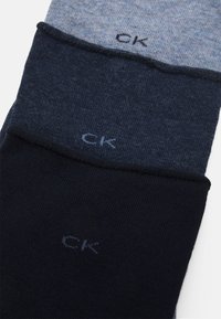 Calvin Klein zeķes trijās krāsās: gaiši pelēka, tumši pelēka un tumši zila. Mīksta auduma, gluda tekstūra, ar izšūtu "CK" logo katrā.