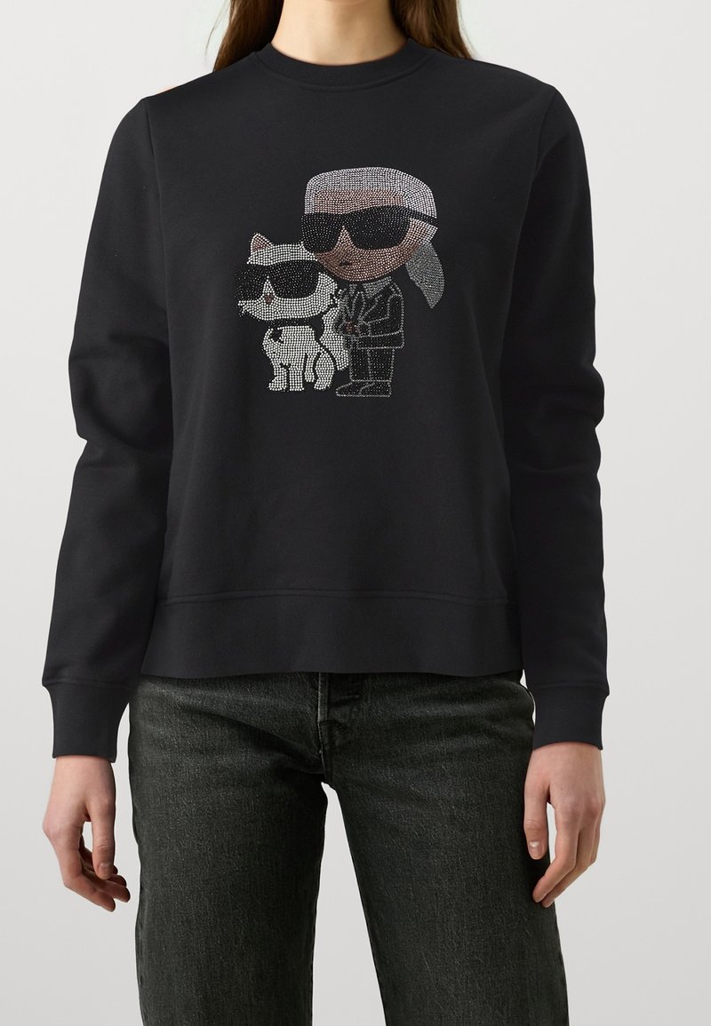 Sweat-shirt noir avec un motif en strass représentant une personne aux cheveux blancs et lunettes de soleil, debout à côté d'un chat portant des lunettes de soleil.