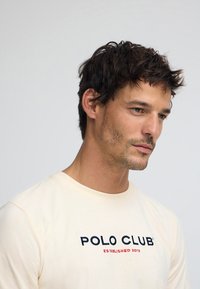 Beige långärmad t-shirt med broderad "POLO CLUB"-logotyp i svart och rött. Mjuk bomullsmaterial, rund halsringning och avslappnad passform.