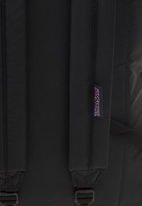 Črn nahrbtnik z oblazinjenimi naramnicami, ojačanimi šivi in vidno oznako Jansport. Narejen iz trpežne tkanine z teksturiranim zaključkom.