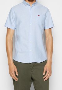 Camicia azzurra a maniche corte con bottoni in tessuto di cotone, dotata di colletto strutturato e taschino sul petto con un piccolo logo di corona rossa.