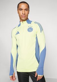 Maglietta atletica gialla chiara con accenti blu, colletto con zip fino a un quarto, maniche lunghe e un logo sul petto, realizzata in un tessuto sportivo e traspirante.