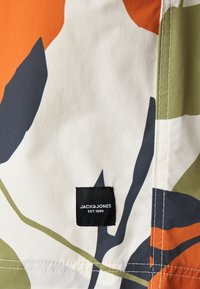 Gemusterter Stoff mit großen abstrakten Formen in Orange, Grün, Dunkelblau und Weiß. Inklusive eines schwarzen Etiketts mit dem Markennamen.
