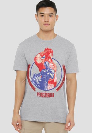 Young man wears gray T-shirt with red and blue Peacemaker comic character illustration and text on front.