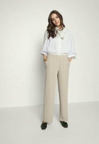 Calças largas beje com cintura elástica, combinadas com uma blusa branca solta e um lenço estampado, usadas com sapatos pretos tipo slip-on.