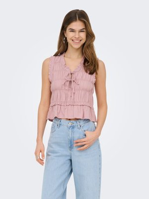 Jonge vrouw die lacht en een mouwloze roze top met ruches draagt en een lichtblauwe spijkerbroek met hoge taille, met één hand in haar zak.