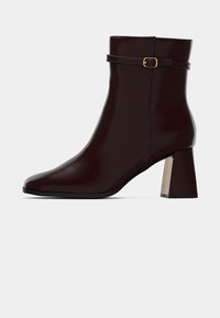 Ciemnobrązowy botek za kostkę na blokowym obcasie z cienkim paskiem z małą złotą klamrą w okolicy górnej części kostki.