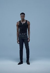 Top preto sem mangas, jeans escuros de corte justo e botins pretos. Colar de corrente prateada. Fundo claro simples realça os detalhes da roupa.